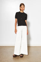 Woman wearing a black short-sleeve top and white wide-leg pants against a plain background