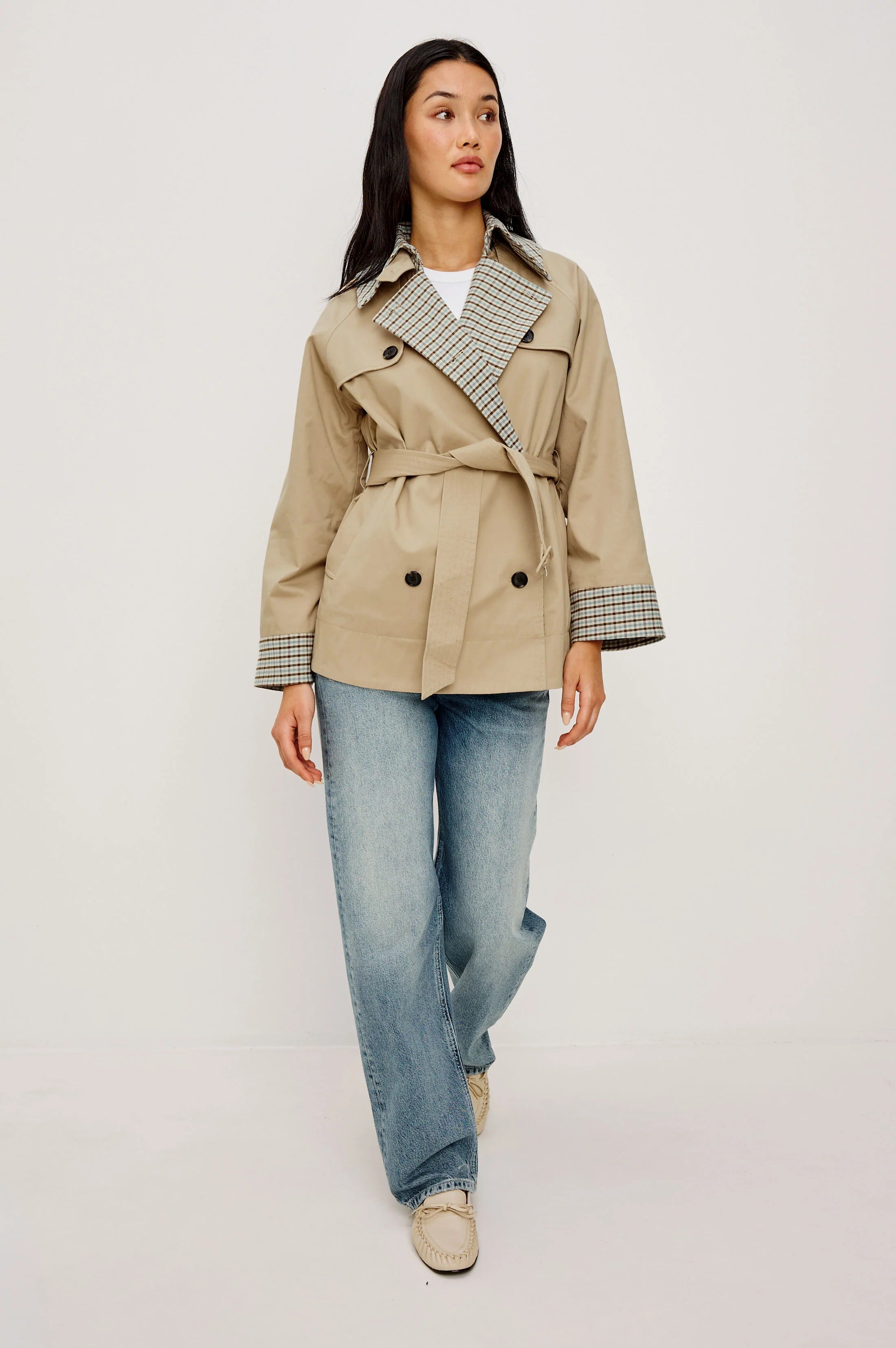 Woman wearing a beige trench coat with plaid accents on a plain background
