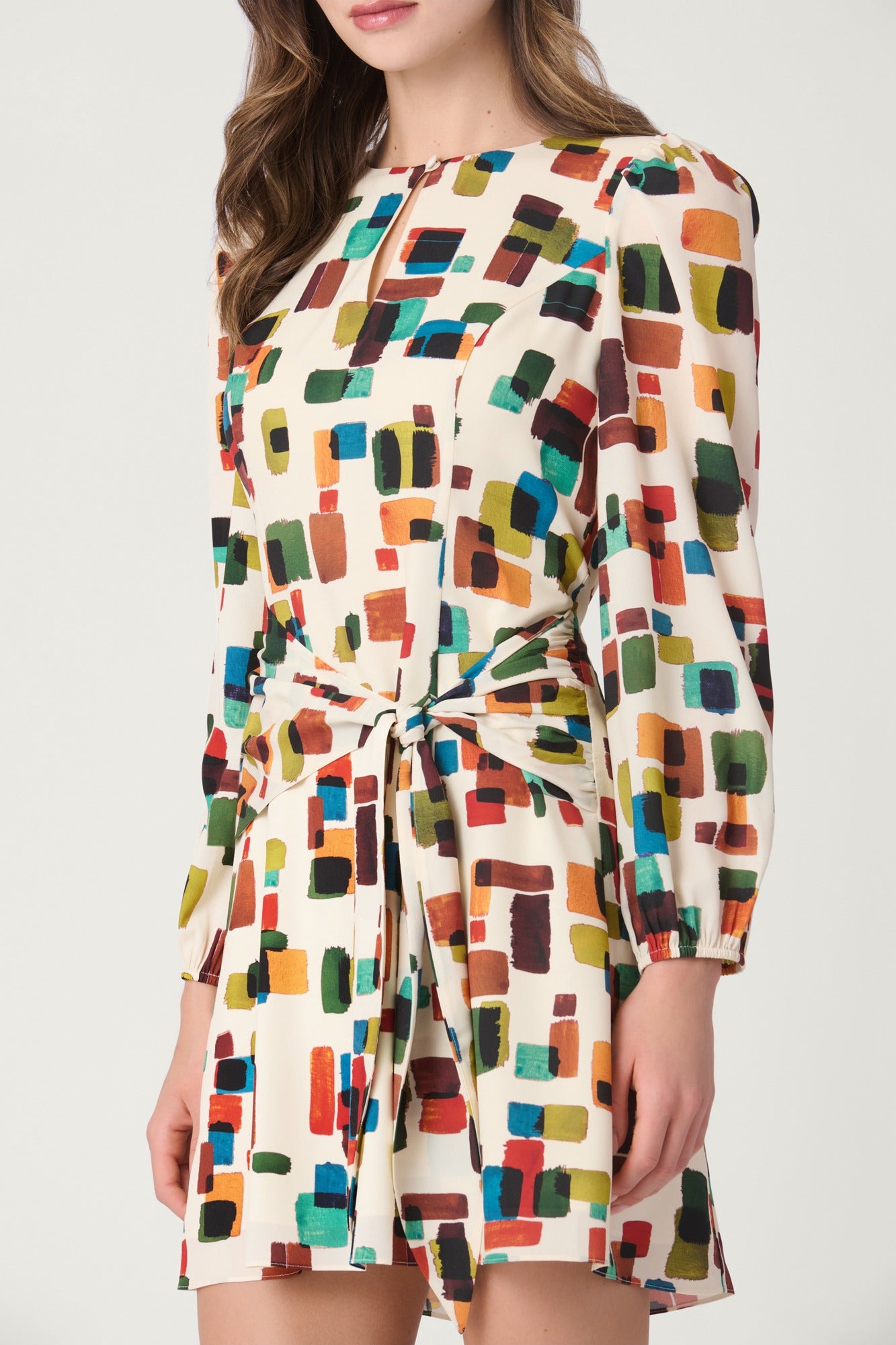 A woman wearing a long-sleeve cream-colored mini dress with a colorful abstract geometric block pattern and a matching waist tie, shown against a plain light gray studio background.