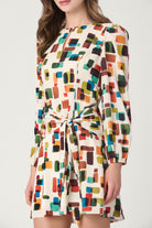 A woman wearing a long-sleeve cream-colored mini dress with a colorful abstract geometric block pattern and a matching waist tie, shown against a plain light gray studio background.
