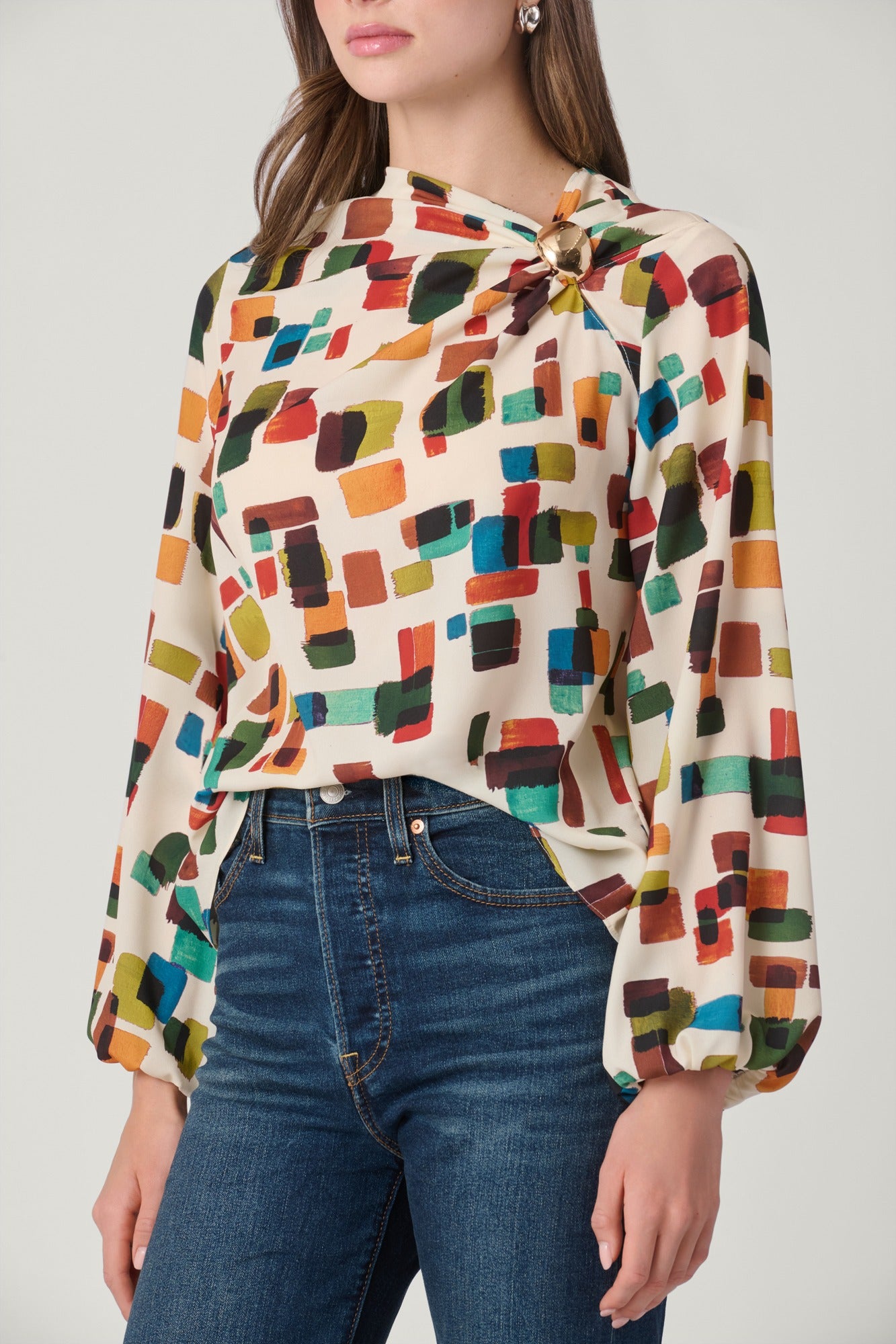 A woman wearing a long-sleeve blouse with a colorful abstract geometric print on a cream background, paired with dark blue jeans, against a light gray studio background.