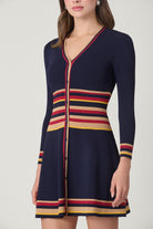 A woman models a navy blue knit cardigan dress with horizontal stripes in red, yellow, and beige, featuring a button-front V-neckline and long sleeves.