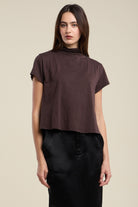 A woman modeling a dark brown short-sleeve mock neck crop top paired with black pants, set against a solid light beige background.