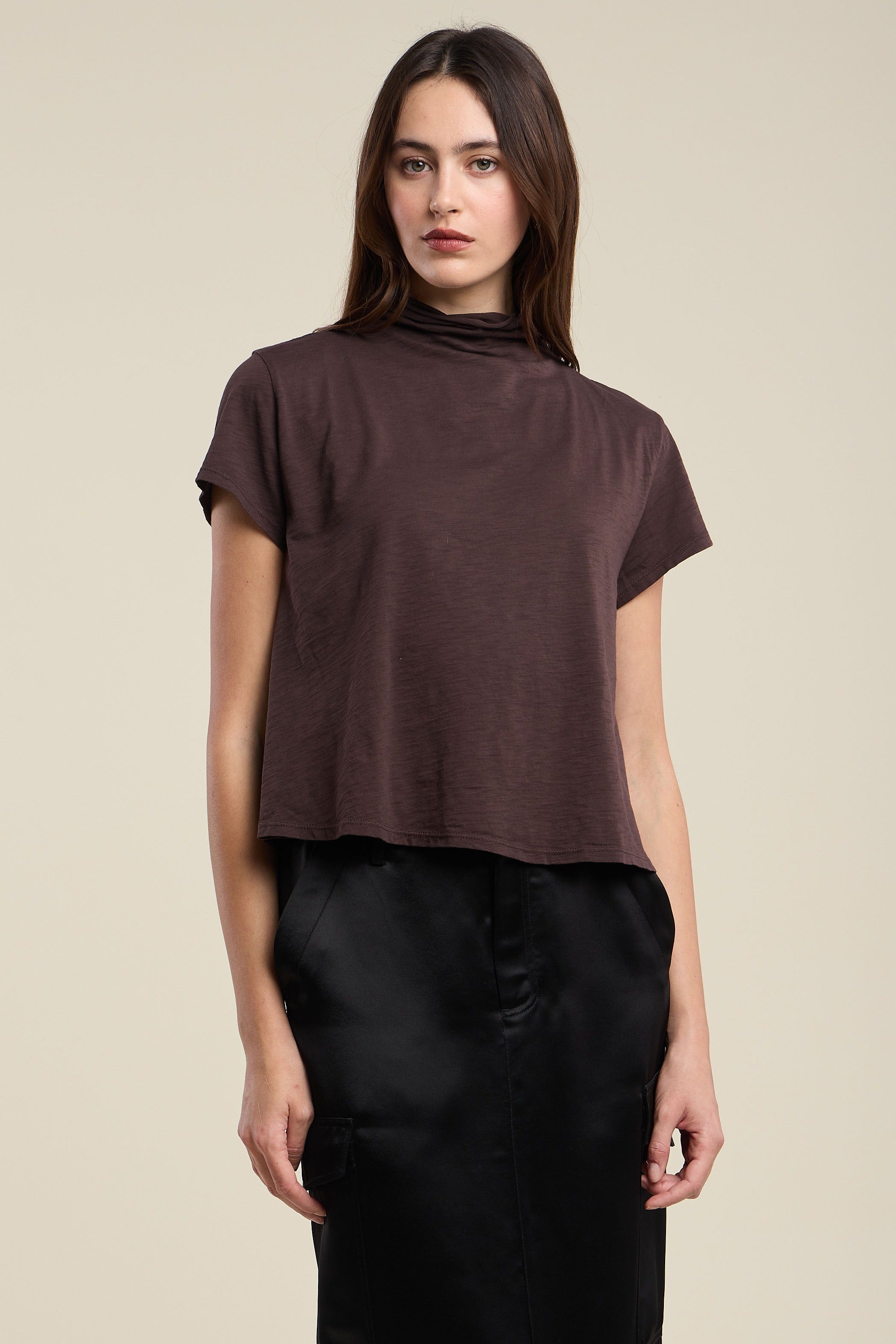 A woman modeling a dark brown short-sleeve mock neck crop top paired with black pants, set against a solid light beige background.