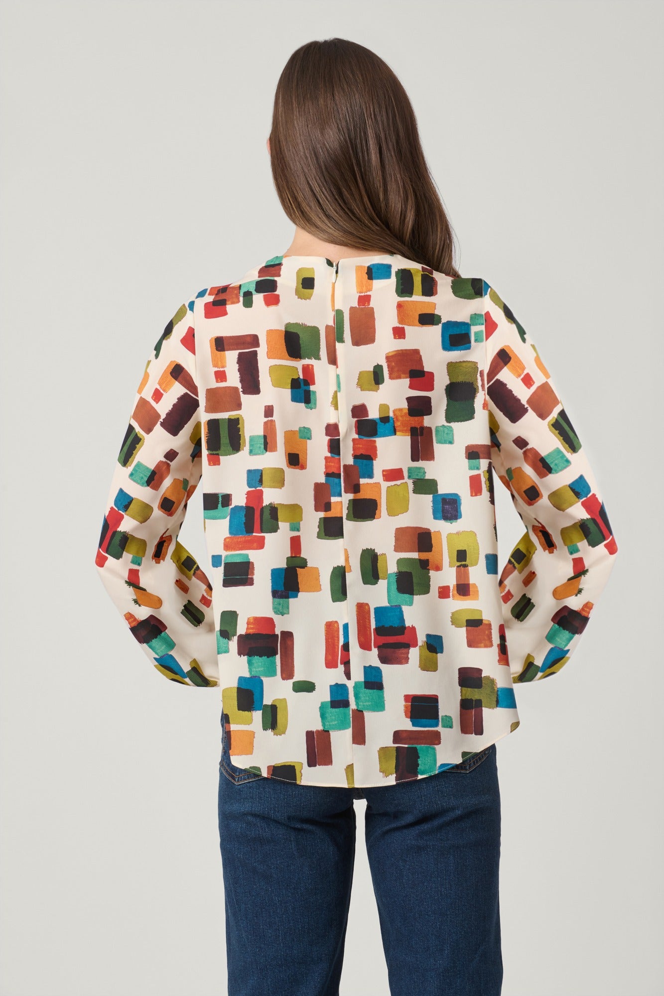 A woman viewed from behind wearing a cream-colored blouse with a colorful geometric block pattern and dark blue jeans, set against a plain light gray studio background.