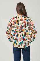 A woman viewed from behind wearing a cream-colored blouse with a colorful geometric block pattern and dark blue jeans, set against a plain light gray studio background.