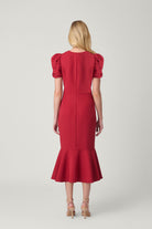 A woman viewed from the back wearing a bright red knee-length dress with short puff sleeves, a straight back seam, and a flared ruffle hem, paired with beige high-heeled sandals against a light gray studio background.