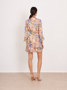 A woman viewed from behind wearing a long-sleeve, ruffled mini dress with a dense floral print in pink, blue, purple, and beige tones, paired with nude high heels against a plain off-white studio background.