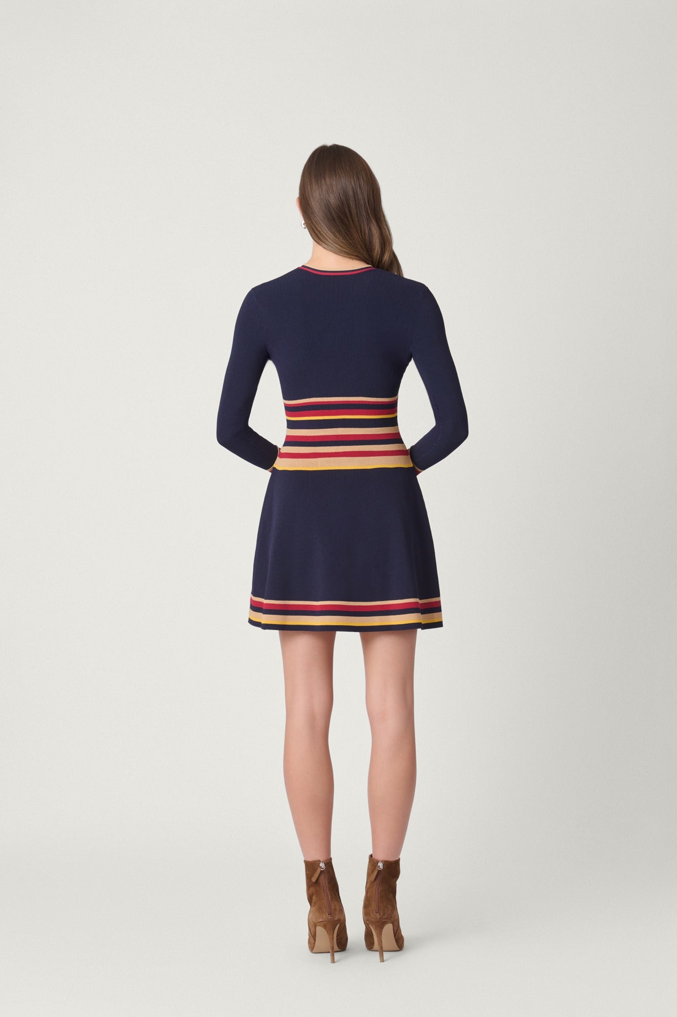 A woman viewed from the back wearing a navy long-sleeve mini dress with horizontal stripes in red, orange, and yellow around the waist and hem, paired with brown ankle boots, against a light gray studio background.
