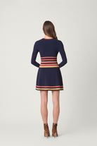 A woman viewed from the back wearing a navy long-sleeve mini dress with horizontal stripes in red, orange, and yellow around the waist and hem, paired with brown ankle boots, against a light gray studio background.