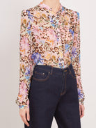 A woman wearing a long-sleeve button-up blouse with a mixed print of pink flowers, blue flowers, and brown leopard spots over a light beige base, paired with dark blue jeans.