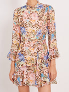 A model wearing a long-sleeve, ruffled mini dress with a busy leopard print and floral pattern in pink, blue, yellow, and brown on a light beige base, set against a plain light gray background.