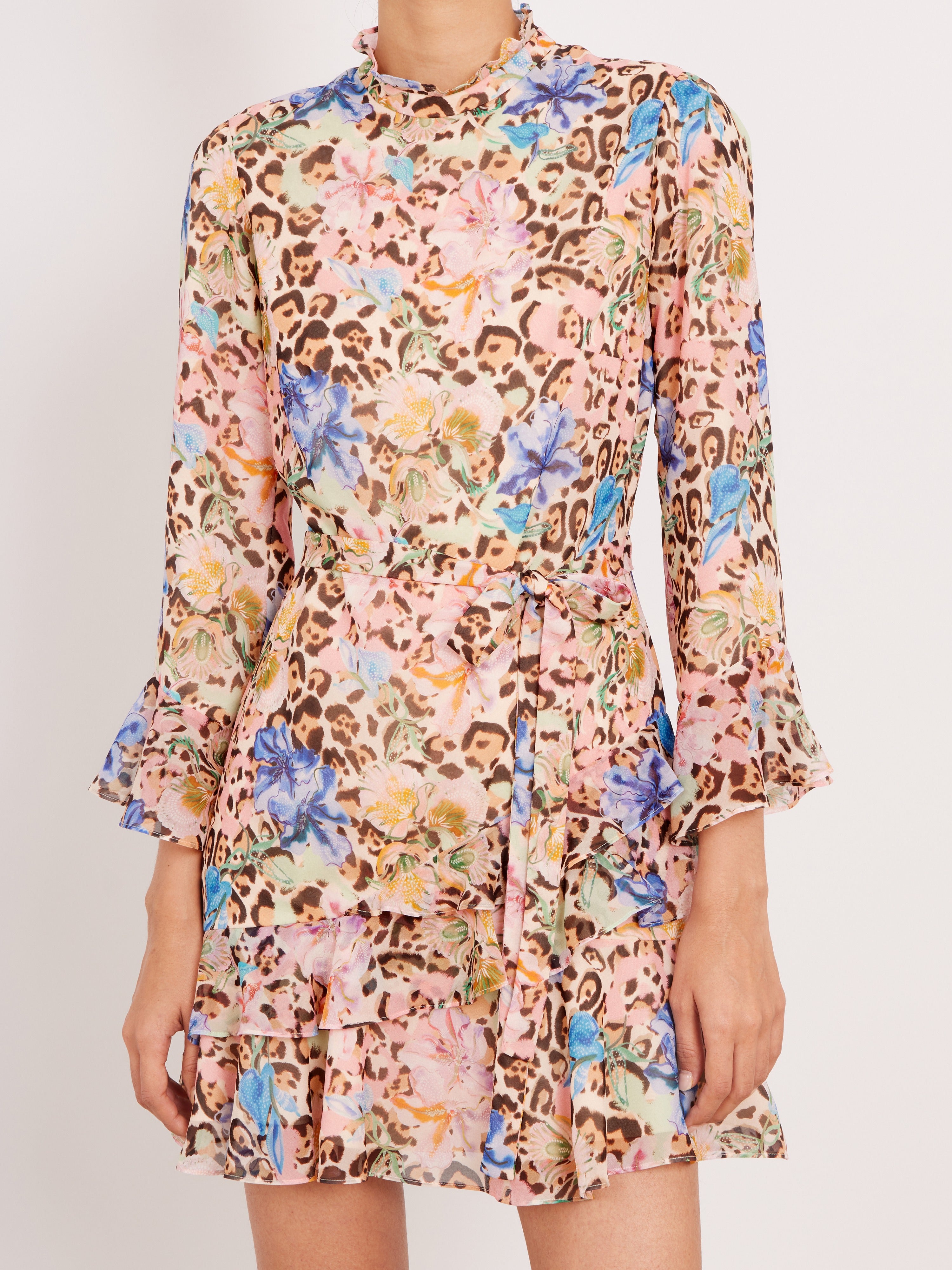 A model wearing a long-sleeve, ruffled mini dress with a busy leopard print and floral pattern in pink, blue, yellow, and brown on a light beige base, set against a plain light gray background.