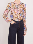 A woman wearing a long-sleeve button-up blouse with a pink, beige, and brown floral leopard print, paired with dark navy wide-leg jeans, against a light gray studio background.