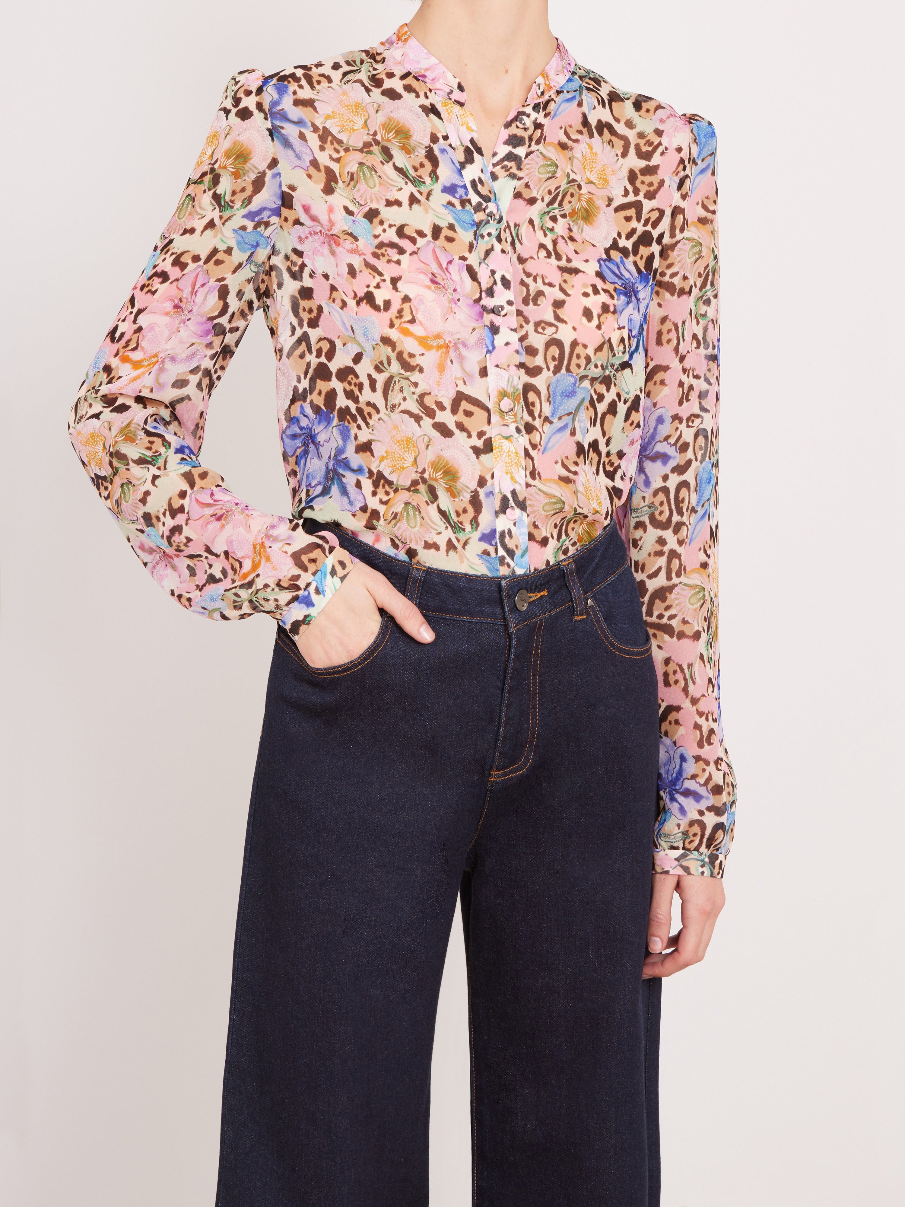 A woman wearing a long-sleeve button-up blouse with a pink, beige, and brown floral leopard print, paired with dark navy wide-leg jeans, against a light gray studio background.