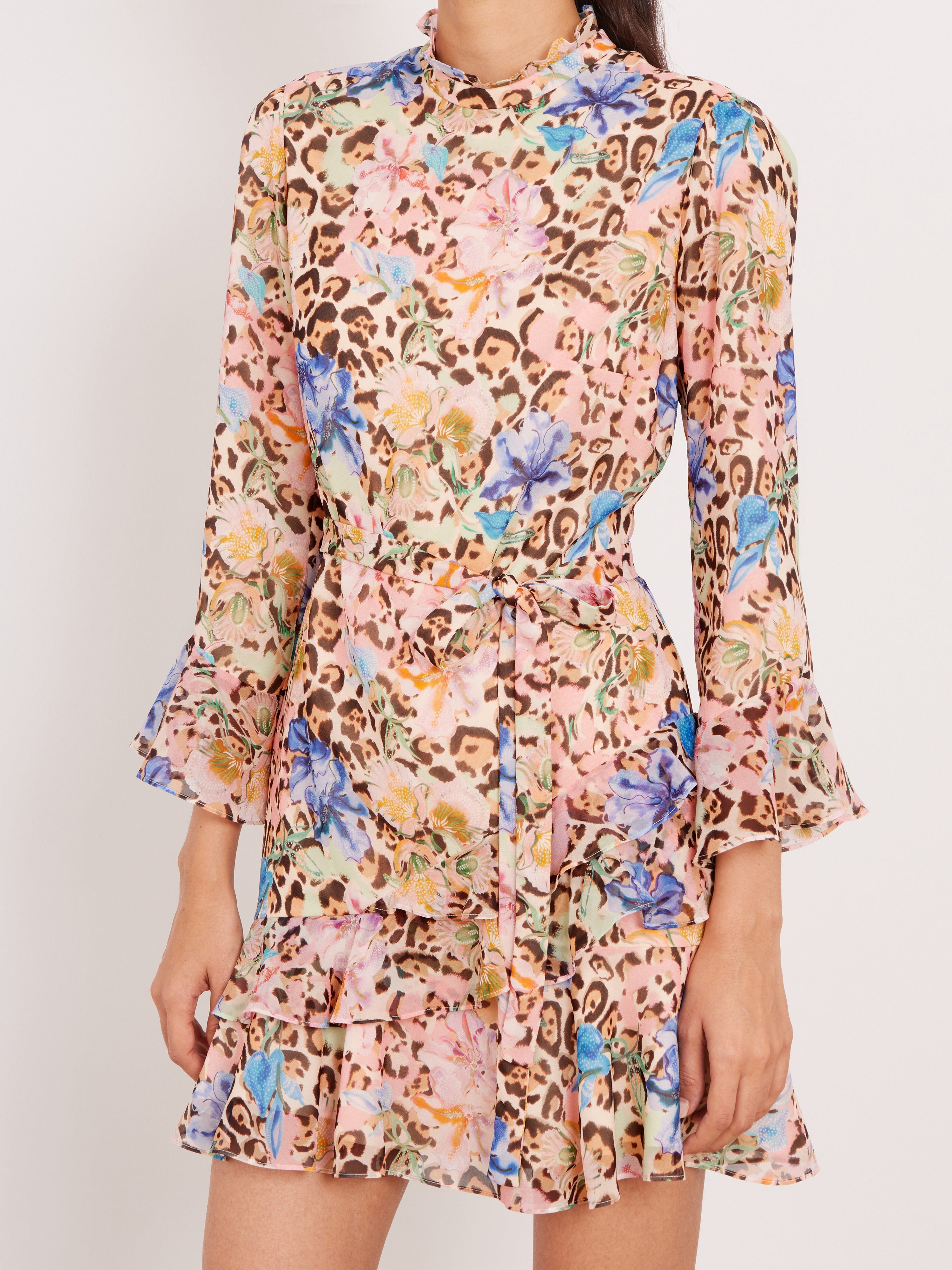 A long-sleeve mini dress with a multicolor floral and leopard print pattern, featuring a high ruffled collar, a self-tie waist belt, and flared bell sleeves, shown on a model against a plain light background.