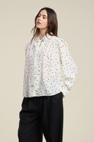 Woman wearing a white blouse with black polka dots and black pants on a beige background