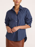 A woman wearing a navy blue button-up shirt with thin white vertical stripes, paired with a brown leather skirt, against a plain light beige background.
