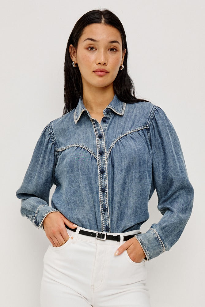 Woman wearing a denim shirt and white pants against a plain background