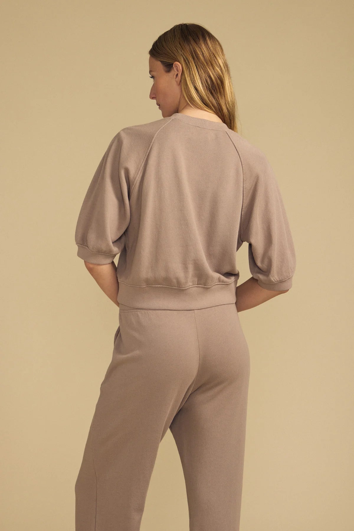 A woman shown from the back wearing a matching taupe lounge set consisting of a short-sleeve cropped sweatshirt and high-waisted pants, set against a solid beige background.