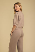 A woman modeling a matching taupe two-piece loungewear set, featuring a short-sleeve cropped sweatshirt with puff sleeves and matching wide-leg pants, set against a solid beige studio background.