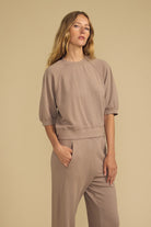 A woman modeling a matching taupe short-sleeve crewneck sweatshirt and wide-leg sweatpants set against a solid beige studio background.