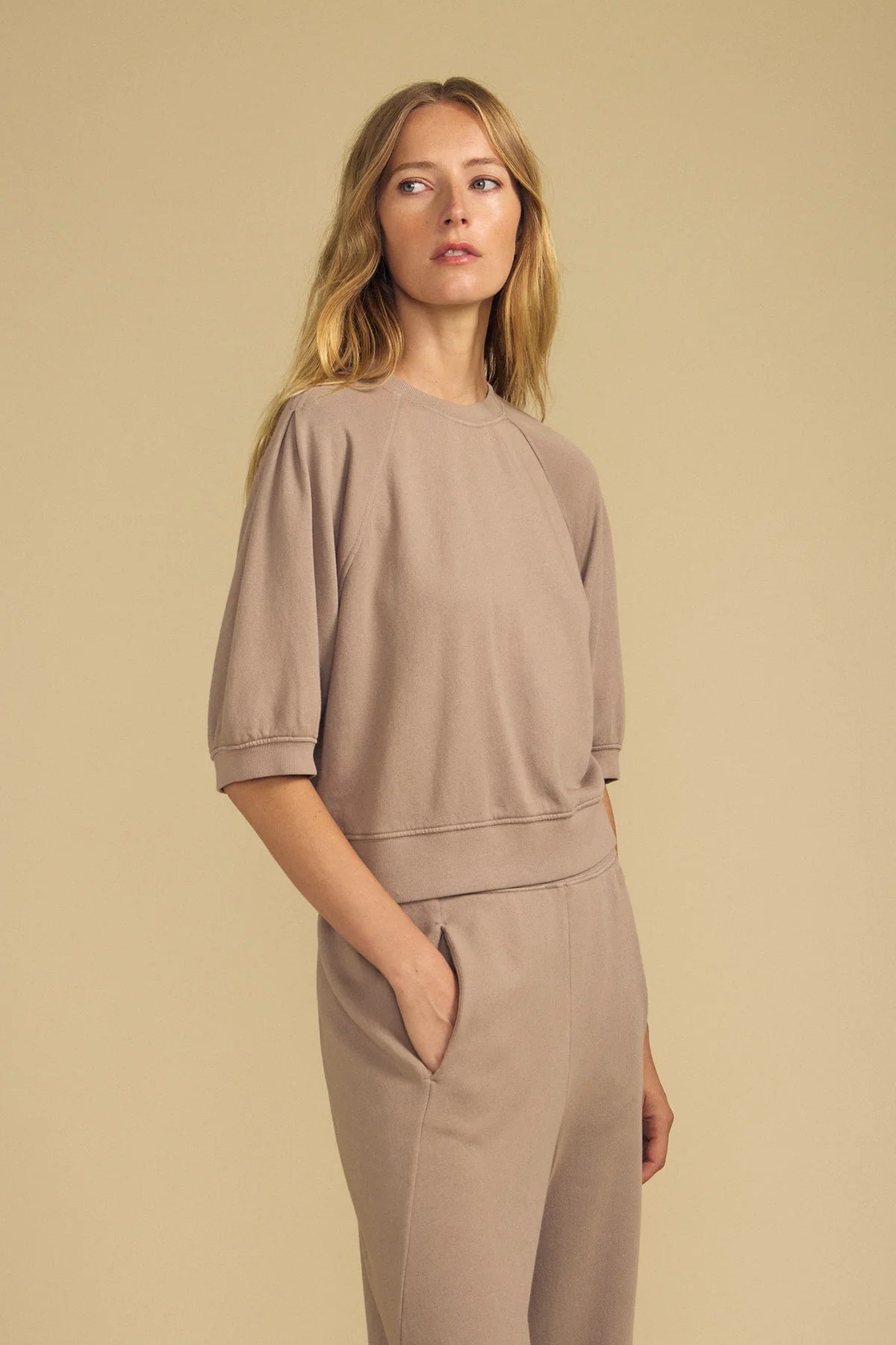 A woman modeling a matching taupe lounge set consisting of a short-sleeve top with gathered shoulders and matching wide-leg pants, set against a solid beige background.