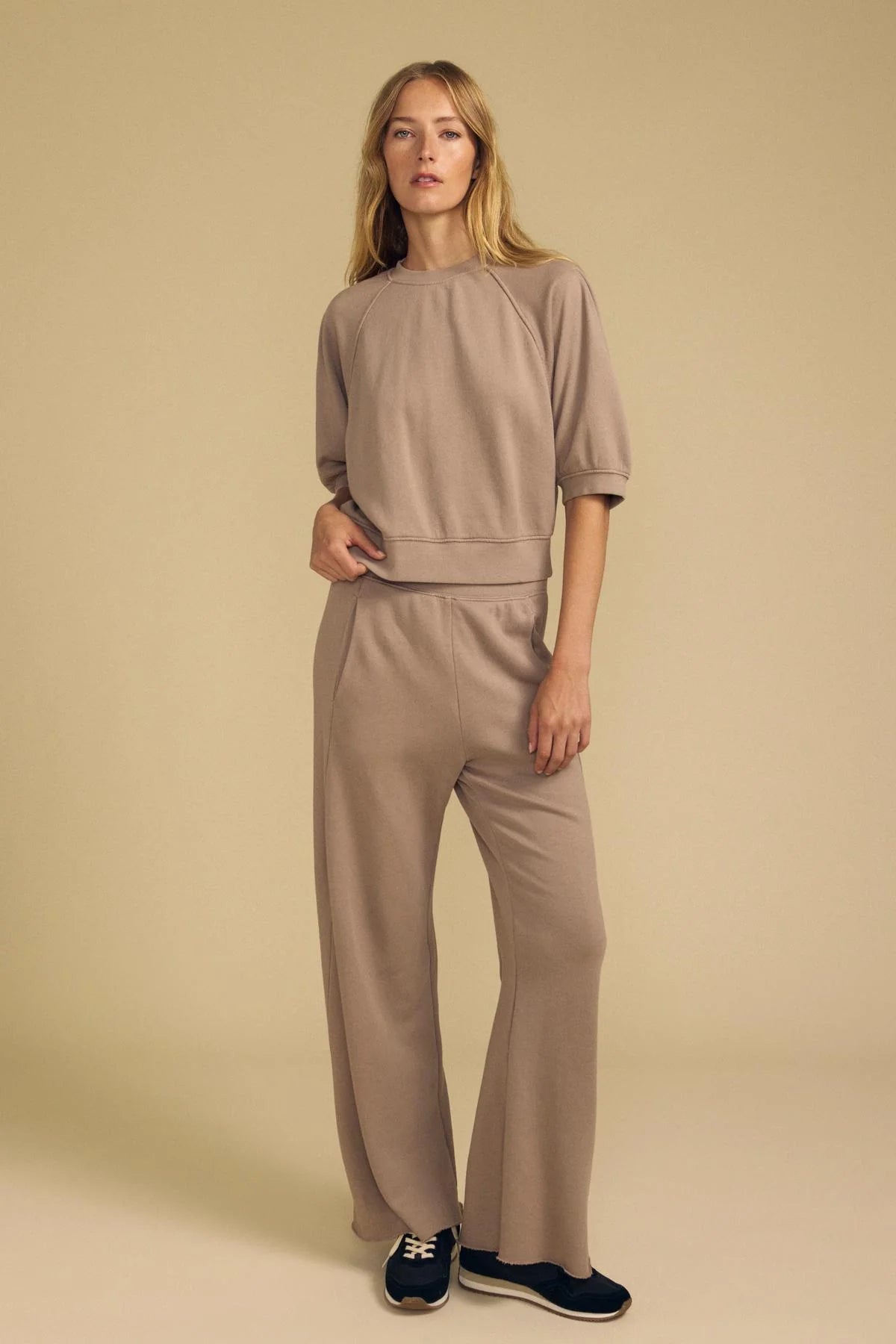 A woman modeling a matching taupe two-piece lounge set consisting of a short-sleeve top and wide-leg pants, paired with navy and white sneakers, against a solid beige studio background.