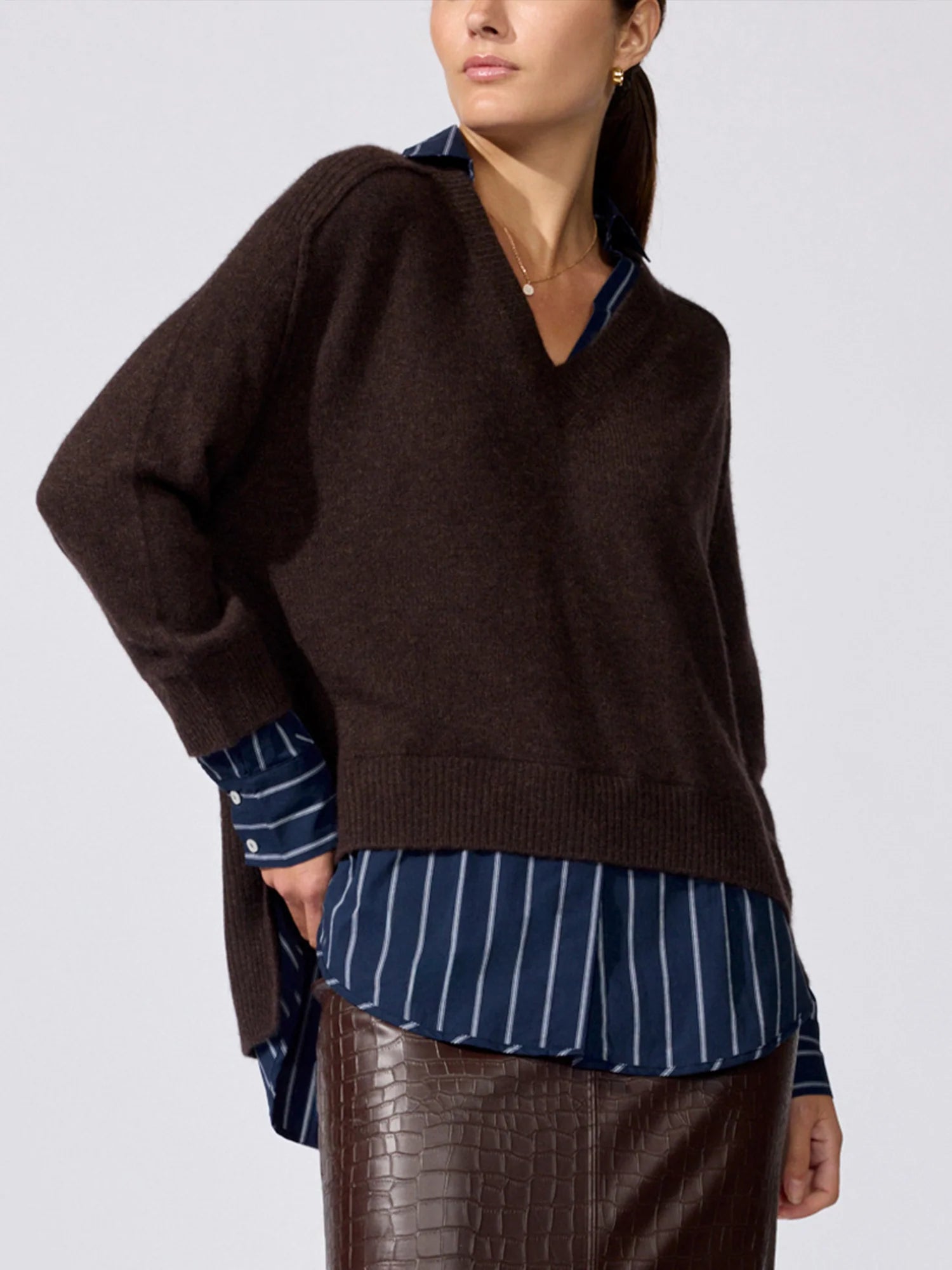 A woman wearing a dark brown knit sweater over a navy blue and white striped shirt, paired with a brown croc-embossed skirt, against a light gray studio background.