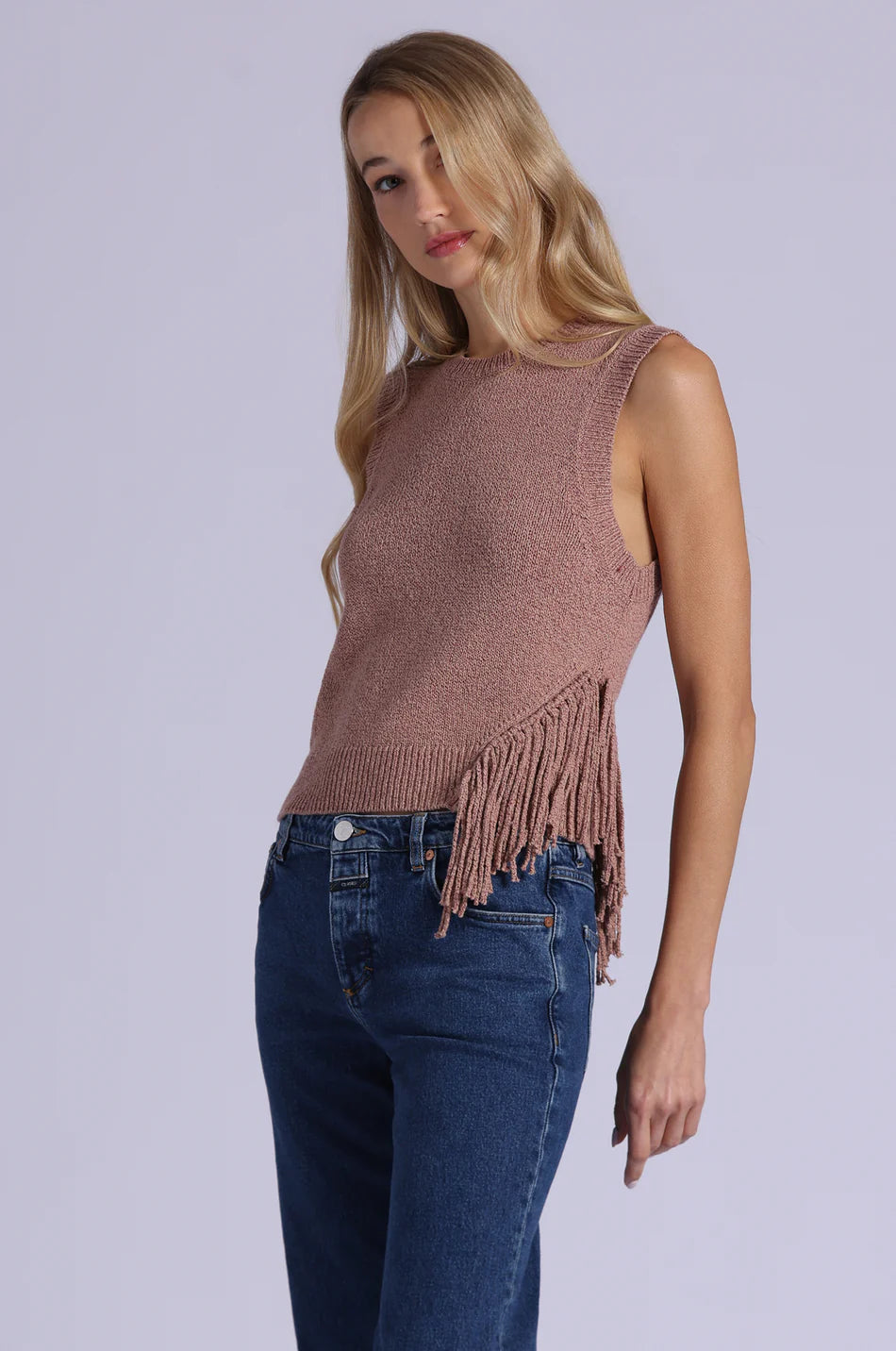 Woman wearing a brown knitted sleeveless top with fringe and blue jeans on a light gray background