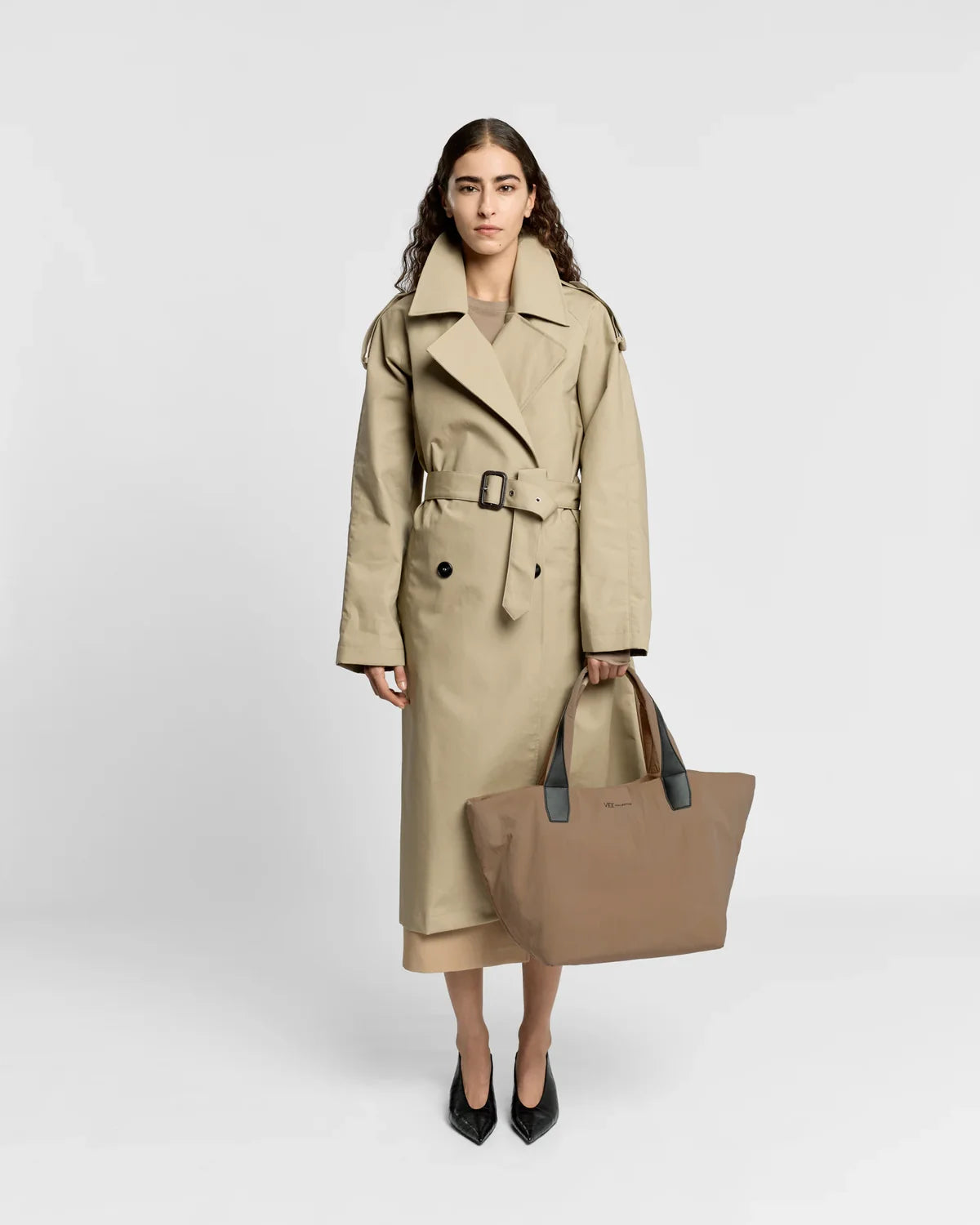 Woman wearing a beige trench coat holding a matching tote bag on a white background