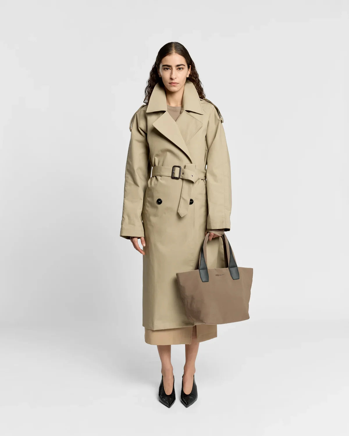 Woman wearing a beige trench coat holding a matching tote bag on a white background