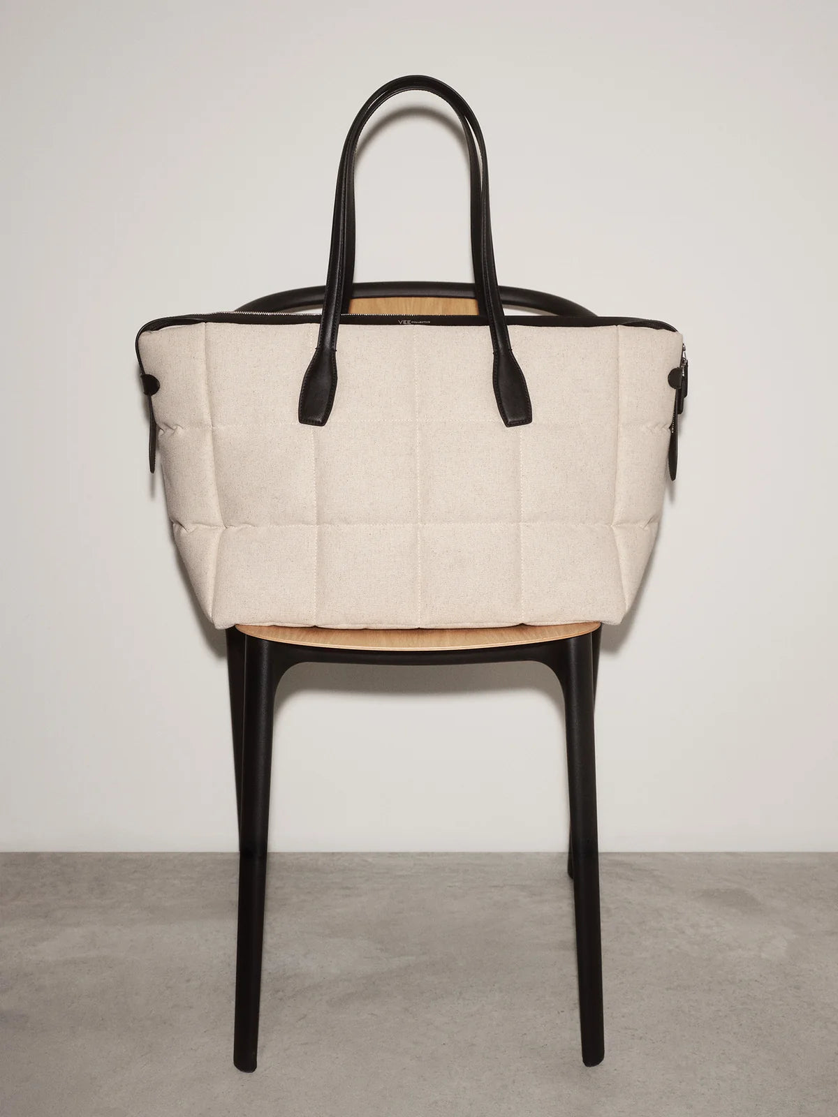 Beige quilted handbag with black handles on a black chair against a white background