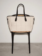 Beige quilted handbag with black handles on a black chair against a white background