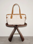 White quilted handbag with brown leather handles and trim on a wooden stand against a light background