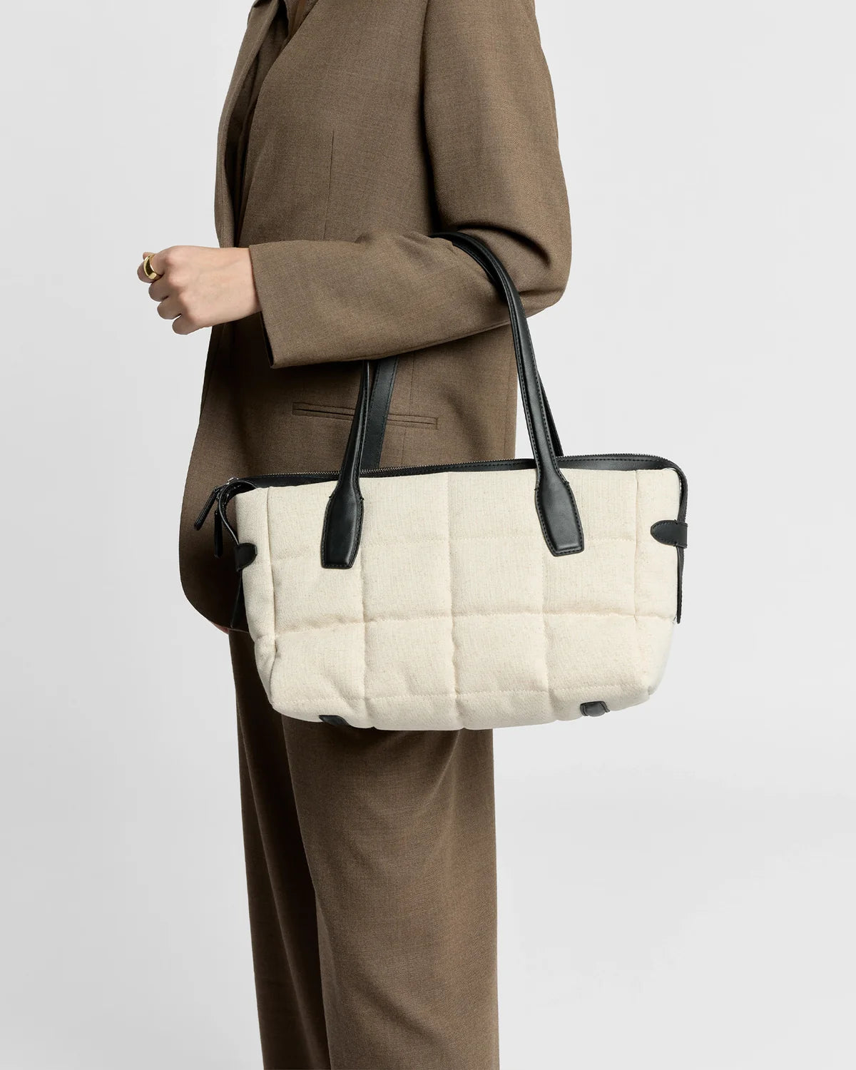 Person wearing a brown coat holding a beige quilted bag with black handles on a light gray background