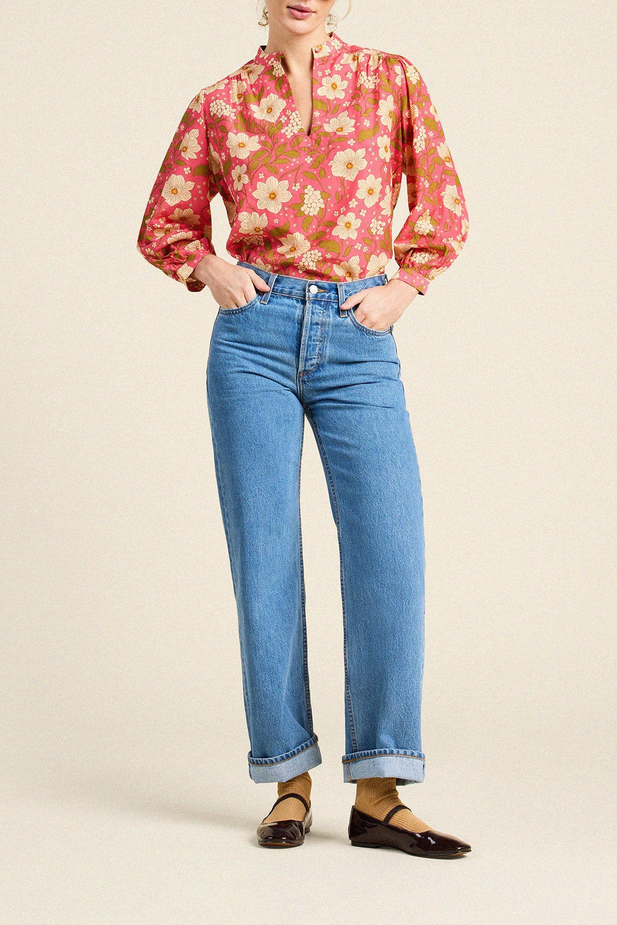 A model wearing a pink and orange floral long-sleeve blouse tucked into light-wash blue jeans, paired with dark brown Mary Jane flats, against a solid beige studio background.