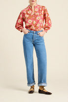 A model wearing a pink and orange floral long-sleeve blouse tucked into light-wash blue jeans, paired with dark brown Mary Jane flats, against a solid beige studio background.