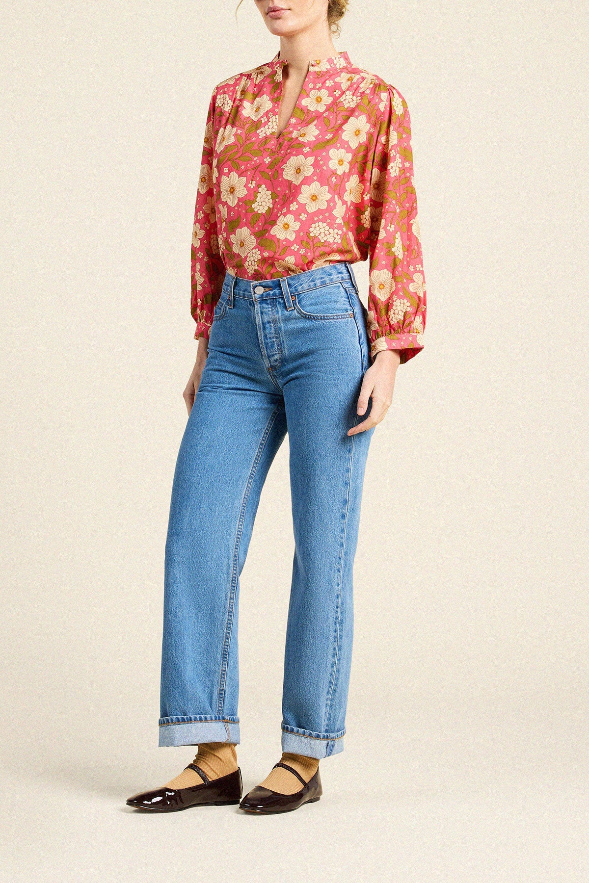 A model wearing a long-sleeve, vibrant red-orange floral blouse with yellow and white flowers, paired with light-wash blue jeans and dark brown flat shoes, set against a solid beige background.