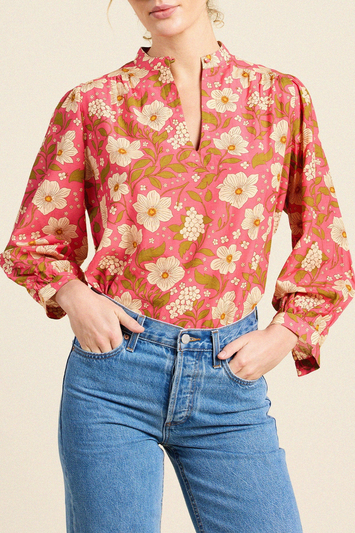 A woman wearing a pink floral long-sleeve blouse with a split neckline and blue jeans, cropped from the mouth to the hips against a plain light beige studio background.