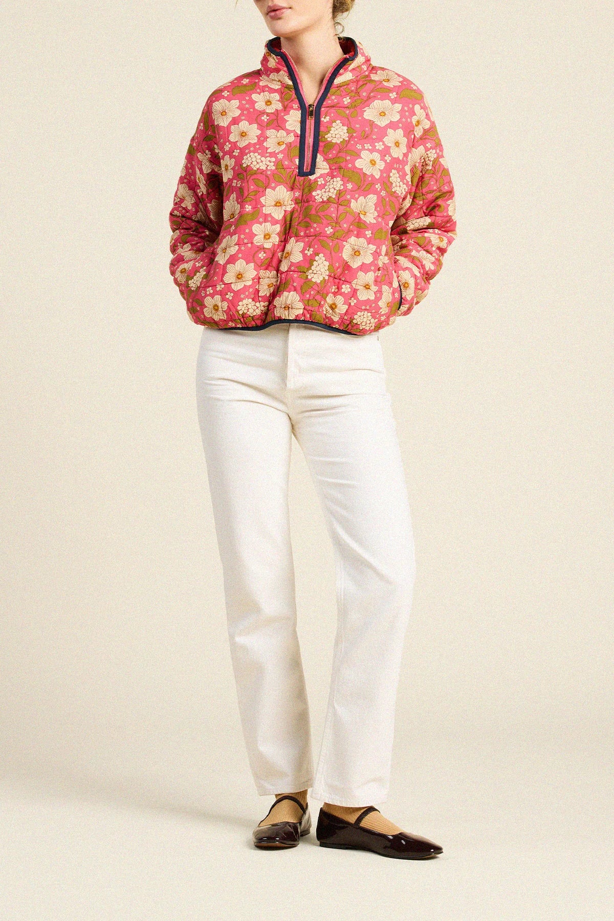 A model wearing a pink floral quarter-zip pullover jacket, white trousers, and dark brown leather flats, standing against a plain beige studio background.