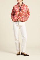 A model wearing a pink floral quarter-zip pullover jacket, white trousers, and dark brown leather flats, standing against a plain beige studio background.