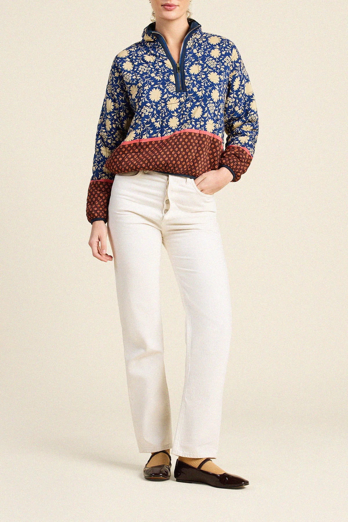 A model wearing a blue and white floral quarter-zip pullover with a rust-brown patterned hem and cuffs, paired with off-white jeans and dark brown flats, against a solid beige background.