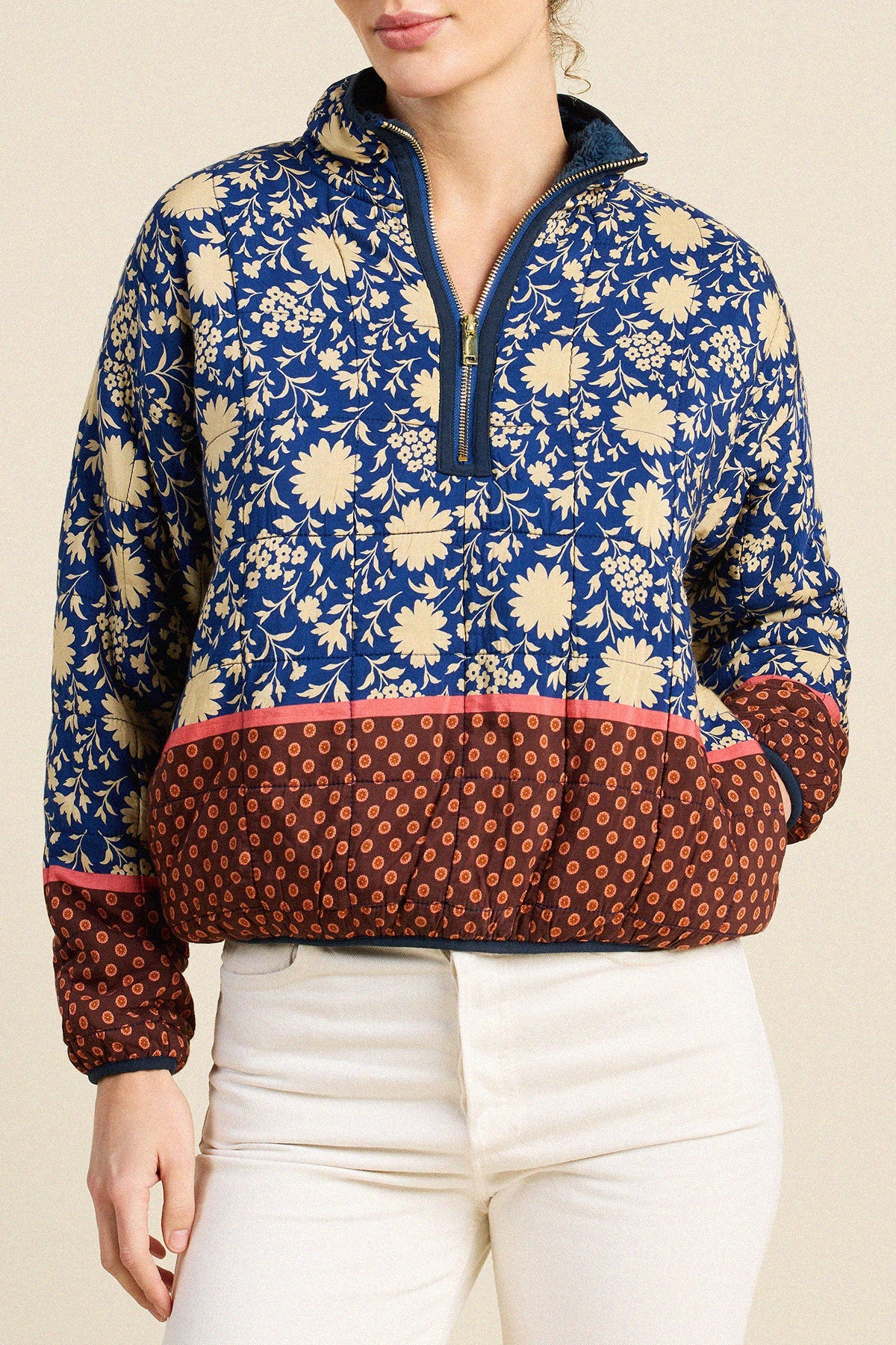 A woman wearing a quarter-zip pullover sweater with a blue and cream floral pattern on the upper body, a solid navy collar and zipper trim, and a rust-brown lower section with a small repeating pattern. She is wearing white pants against a plain beige background.