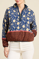 A woman wearing a quarter-zip pullover sweater with a blue and cream floral pattern on the upper body, a solid navy collar and zipper trim, and a rust-brown lower section with a small repeating pattern. She is wearing white pants against a plain beige background.