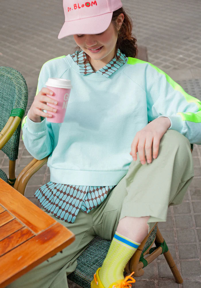 Person sitting outdoors holding a pink cup, wearing a light blue sweater and green pants.
