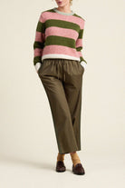 A model wearing a pink and olive green striped knit sweater, olive green wide-leg cropped pants, and dark brown loafers, standing against a plain beige studio background.