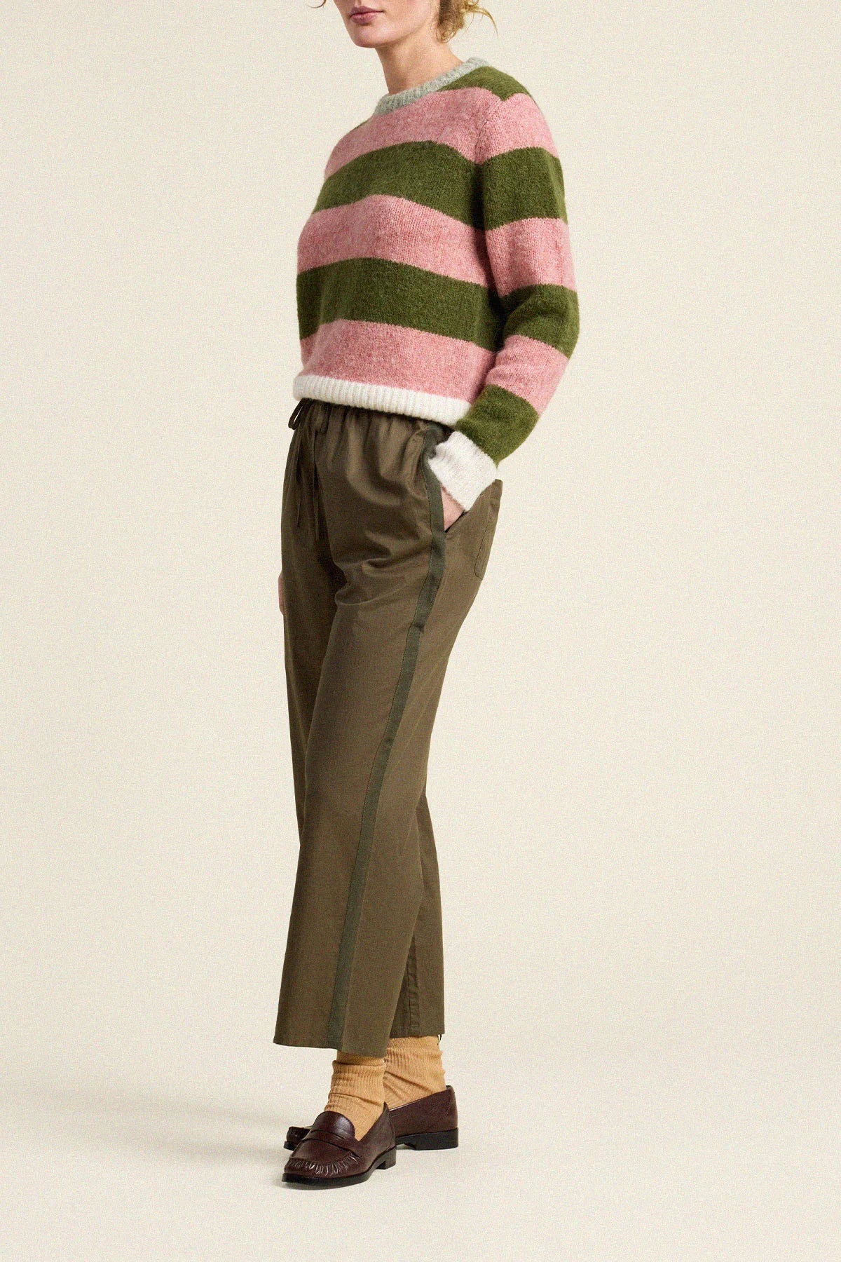 A model is shown from the side wearing a fuzzy knit sweater with horizontal pink and olive green stripes, paired with olive green trousers, tan socks, and dark brown loafers, against a plain beige background.