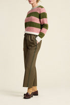 A model is shown from the side wearing a fuzzy knit sweater with horizontal pink and olive green stripes, paired with olive green trousers, tan socks, and dark brown loafers, against a plain beige background.