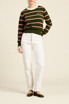 A model wearing a green and pink striped knit sweater, white trousers, and dark brown leather flats, standing against a solid beige background.
